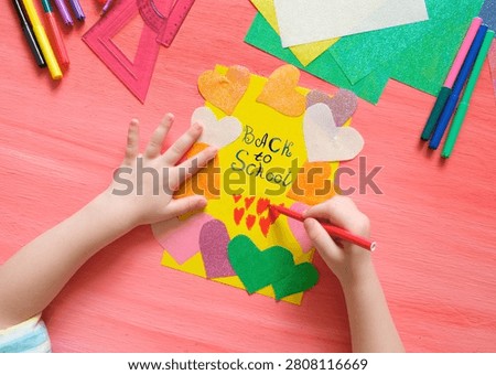 Back to School’ paintbrush text.  Child getting ready to create a greeting card.  On the table typical crafting materials. Showcases creativity, DIY, and craft activities. School supplies, art projects, educational content. Educational content about arts and crafts.