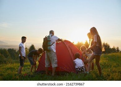 Back Panoramic View Of Group Of Children Together With Teacher Preparing Campsite In Mountains. Kids With Old Man Rucksack Setting Up Red Tent At Sunset. Concept Of Hiking.