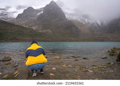 Back Of A Person Crouching On The Shore Of A Lake With Snowy Peaks On The Background. Chacas, Peru