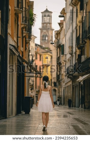 The back of a petit Asian girl in a white dress walking down a rustic empty side street in the Italian town of Verona on a sunny summers day