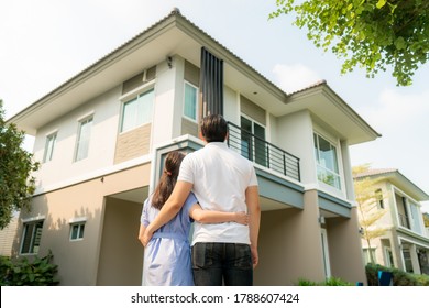 Back portrait of Asian young couple standing and hugging together looking happy in front of their new house to start new life. Family, age, home, real estate and people concept.