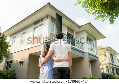 Back portrait of Asian young couple standing and hugging together looking happy in front of their new house to start new life. Family, age, home, real estate and people concept.