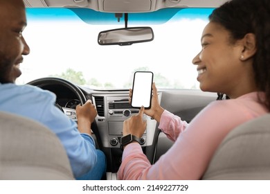 Back rear over the shoulder view of happy black female passenger holding cell in hand pointing finger at white blank mock up screen, sitting on front seat using mobile application for city navigation