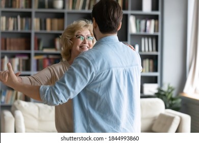 Back Rear View Affectionate Caring Young Man Leading In Slow Dance Smiling Middle Aged 60s Senior Mother At Home. Happy Proud Loving Old Mature Woman Enjoying Waltz With Grownup Son In Living Room.