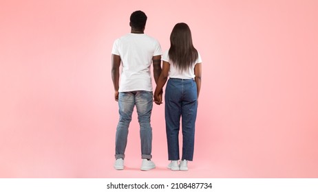 Back Rear View Of Casual Black Young Couple Holding Hands And Looking At Empty Wall Together, Guy And Lady Posing Isolated On Pink Studio Background, Full Body Length, Panorama Banner, Free Copy Space