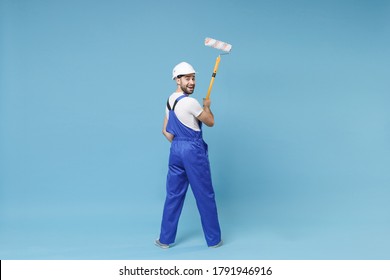 Back rear view of cheerful young man in coveralls protective helmet hardhat hold paint roller isolated on blue background. Instruments accessories for renovation apartment room. Repair home concept