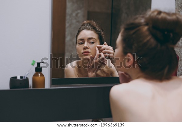 Back rear view disappointed half naked topless\
young woman 20s playing with her face squeeze pimple reflected in\
mirror in bathroom do morning routine Skin care healthcare cosmetic\
procedures concept