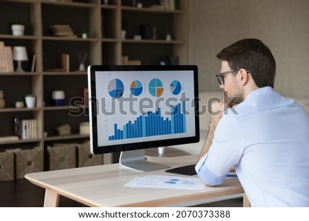 Back rear view focused young businessman looking at computer monitor, analyzing project statistics, marketing research results or statistics data, developing marketing strategy, working at home office