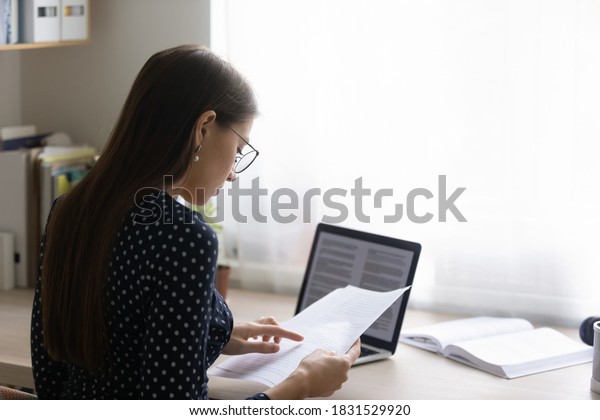 Back rear view focused young woman in eyeglasses\
reading paper document, checking electronic report on computer.\
Concentrated smart ambitious businesswoman preparing research,\
freelance work.