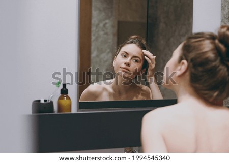 Back rear view nice half naked topless caucasian young woman 20s touching forehead reflected in mirror in bathroom doing morning routine. Skin care healthcare cosmetic procedures lifestyle concept.