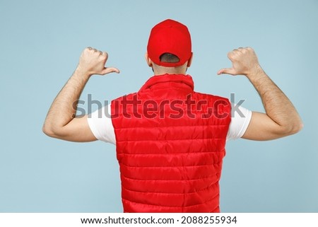 Back rear view of professional delivery guy employee man in red cap white T-shirt vest uniform workwear work as dealer courier isolated on pastel blue color background studio portrait. Service concept