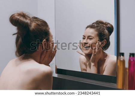 Back rear view smiling half naked topless young woman 20s in bra look at face reflected in mirror touch face in bathroom do morning routine bodycare. Skin care healthcare cosmetic procedures concept