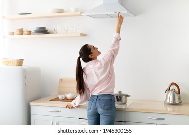 Back rear view of smiling woman selecting mode on cooking hood, standing near kitchen appliance in contemporary interior and decor with shelves, pushing button on mechanical fan above the stove