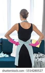 Back Rear View Of Young Adult Woman Holding Hands On Hips, Standing In Living Room And Looking At Mess On Sofa