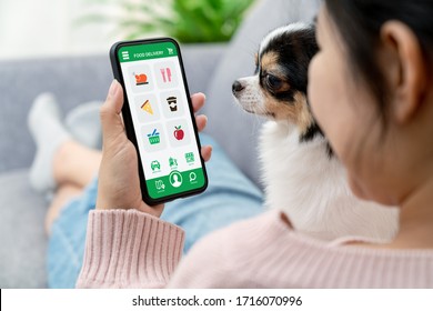 Back rear view of young asian teenage girl in concept next normal life from coronavirus impact sitting at sofa couch with dog hand holding smart phone at home. Food or grocery delivery order service.