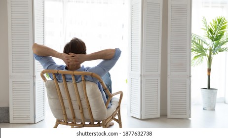 Back rear view young caucasian man relaxing on comfortable chair with folded arms behind hand, looking out of window, contemplating or daydreaming alone in light living room, enjoying peaceful moment.