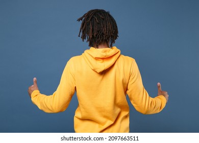 Back Rear View Of Young Man Of African American Ethnicity Wearing Yellow Streetwear Hoodie Posing Isolated On Blue Wall Background Studio Portrait. People Emotions Lifestyle Concept. Showing Thumbs Up