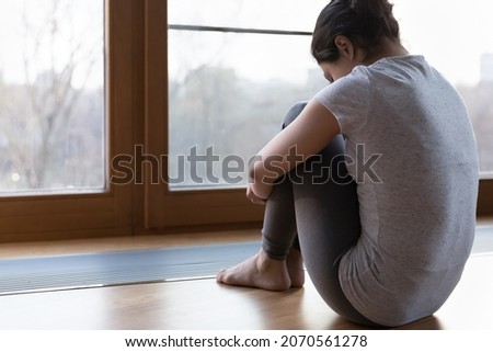 Back rear view young miserable unhappy female emb knees seated on floor near window feels lonely, thinks over life concerns, experiences personal troubles, teenager suffers from bullying concept