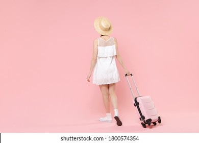 Back rear view young tourist girl in summer white dress hat with suitcase isolated on pink background studio portrait.  
