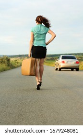 Back, Rear View. Young Woman Walking With Retro Siutcase On A Country Asphalt Road. Girl With Heavy Bag Walking Along Road. From Below Shot Of Pretty Young With Suitcase