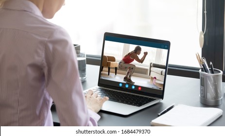 Back Rear View Young Woman Sitting At Home Office Workplace, Watching Educational Fitness Training Video On Laptop. Screen View Healthy Trainer In Sportwear Doing Squats, Presenting Online Course.