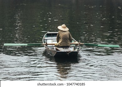 Back of rowing boat man wearing green shirt and conical hat sitting in a boat with paddles over the river in background at Trang An Grottoes in Ninh Binh, Vietnam.