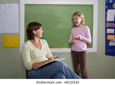 Back To School - 8 Year Old Student And Teacher Talking By Blackboard In Classroom