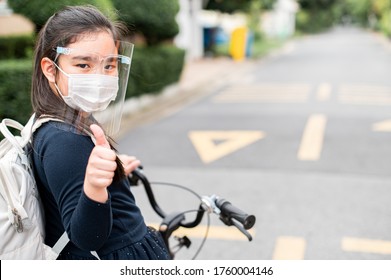 Back To School. Asian Child Girl Wearing Face Mask And Giving Thumb Up With Backpack Biking A Bicycle And Going To School .Covid-19 Coronavirus Pandemic.New Normal Lifestyle.Education Concept.