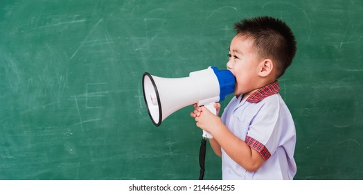 Back To School. Asian Funny Cute Little Child Boy Kindergarten Preschool In Student Uniform Speaking Through Megaphone Against On Green School Blackboard, First Time To School Education Concept