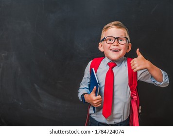 Back to school. Blond hair boy in glasses and school uniform showing thumbs up gesture. Happy pupil with book and bag near blackboard. Successful little businessman - Shutterstock ID 1784724074