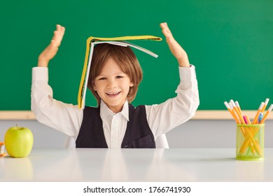 Back To School. Cheerful Little Boy Hiding Under Book, Playing Silly Game At Desk In Classroom.