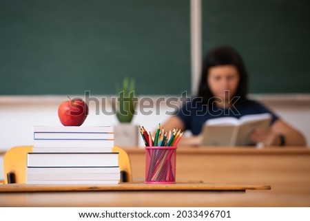 Back to School Concept. Apple, accessories and books in the classroom, chalkboard and teacher teaching in the background.