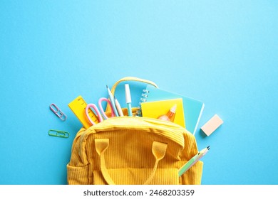 Back to School Concept. Brightly colored yellow backpack overflowing with essential school supplies such as notebooks, pens, pencils, scissors, and a ruler on a vibrant blue background. Flat lay.