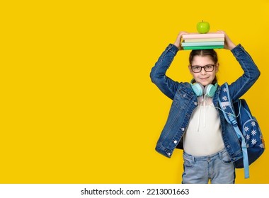 Back To School Concept. Little Happy Girl In Eyeglasses And Headphones With Stack Of Books On Her Head On Yellow Background. A Schoolgirl Holds A Textbooks On Her Head