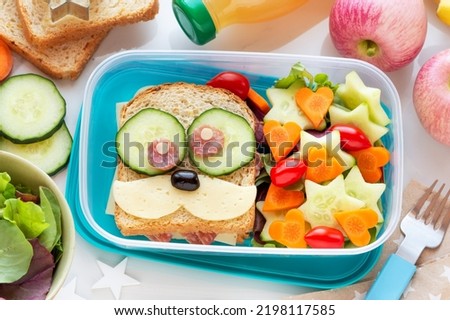 Back to school concept. Lunch box preparation for kids with a cheese and salami sandwich, fresh salad leaves, cucumbers, carrots and tomatoes 
