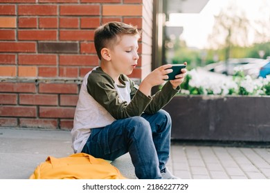 Back To School. Cute Child With Backpack, Holding Mobile Phone, Playing With Cellphone. School Boy Pupil With Bag. Elementary School Student After Classes. Kid Sitting On Stairs Outdoors In The