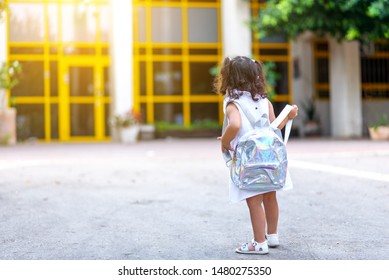 Back to school. Cute child girl with holographic backpack running and going to school with fun.Back, rear view of a toddler kid walking towards the preschool with the door in the background.
