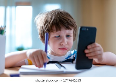 Back To School. Distance Learning Online Education. Caucasian Sad Kid Boy Studying At Home With Smartphone And Doing Homework. Sad Child Siting At Table With Cell Phone.