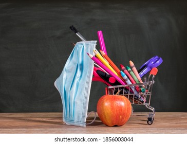 Back To School During Coronavirus Pandemic With Pencils, Crayons And Face Mask In Shopping Cart With Apple Against Chalkboard