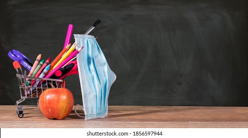 Back To School During Coronavirus Pandemic With Pencils, Crayons And Face Mask In Shopping Cart With Apple Against Chalkboard