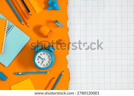 Back to school flat lay background with blue alarm clock, pencils, notebook and colorful stationery on orange torn paper next to blank checkered copy space, ready for your creative educational text.