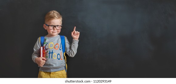 Back to school. Funny little boy in glasses pointing up on blackboard. Child from elementary school with book and bag. Education. - Shutterstock ID 1120293149