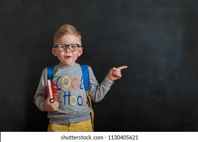 Back To School. Funny Little Boy In Glasses Pointing Up On Blackboard. Child From Elementary School With Book And Bag. Education. Kid With A Book