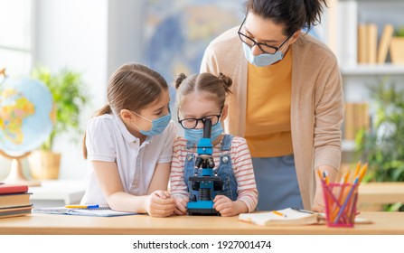 Back to school. Happy children are wearing facemasks sitting at desks. Girls with teacher in classroom. - Shutterstock ID 1927001474