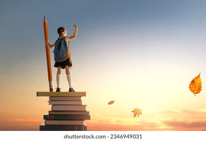Back to school! Happy cute industrious child standing on the tower of books and holding a huge pencil on background of sunset sky. Concept of education and reading. The development of the imagination. - Shutterstock ID 2346949031