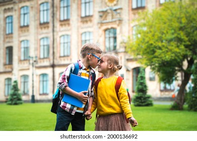 Back to school. Happy little kids hugging and kissing each other. School first love. Boy kissing a girl in nose. Little students with backpacks and notebook in school yard. Friends.