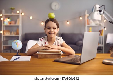 Back To School. Happy Schoolgirl With Apple Ina Head Smiles While Sitting At The Table With Laptop At Home.