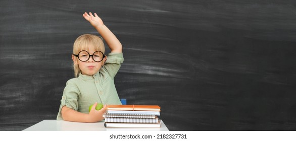 Back To School. Happy Smiling Pupil In Round Glasses Sitting At The Desk. Child Raises His Hand Up In The Classroom With Blackboard On Background. Kid Boy From Primary School. First Day Of Fall.