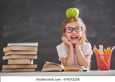 Back To School And Happy Time! Cute Industrious Child Is Sitting At A Desk Indoors. Kid Is Learning In Class On Background Of Blackboard. Girl Reading The Book.