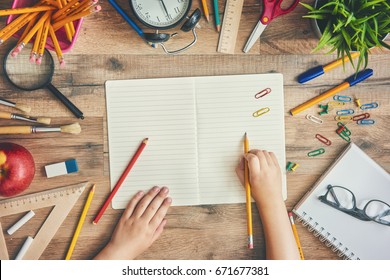 Back To School And Happy Time! Wooden Desk With Supplies. A Child Writes In An Empty Notebook. Top View. 